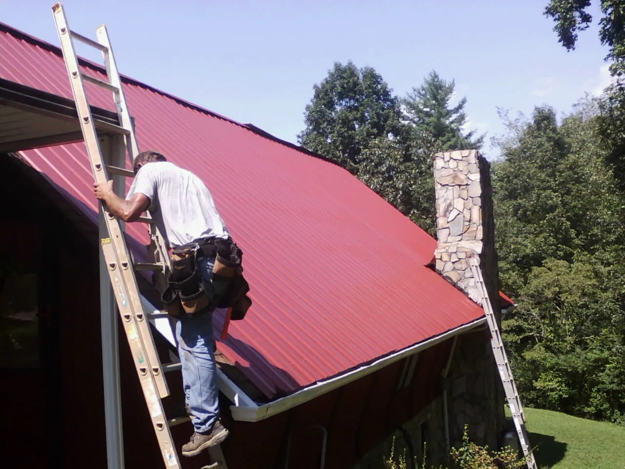 Roofer on a ladder beside a red metal roof and stone chimney — Soffit & Fascia Repair by Roofing Brothers in Rockville Centre